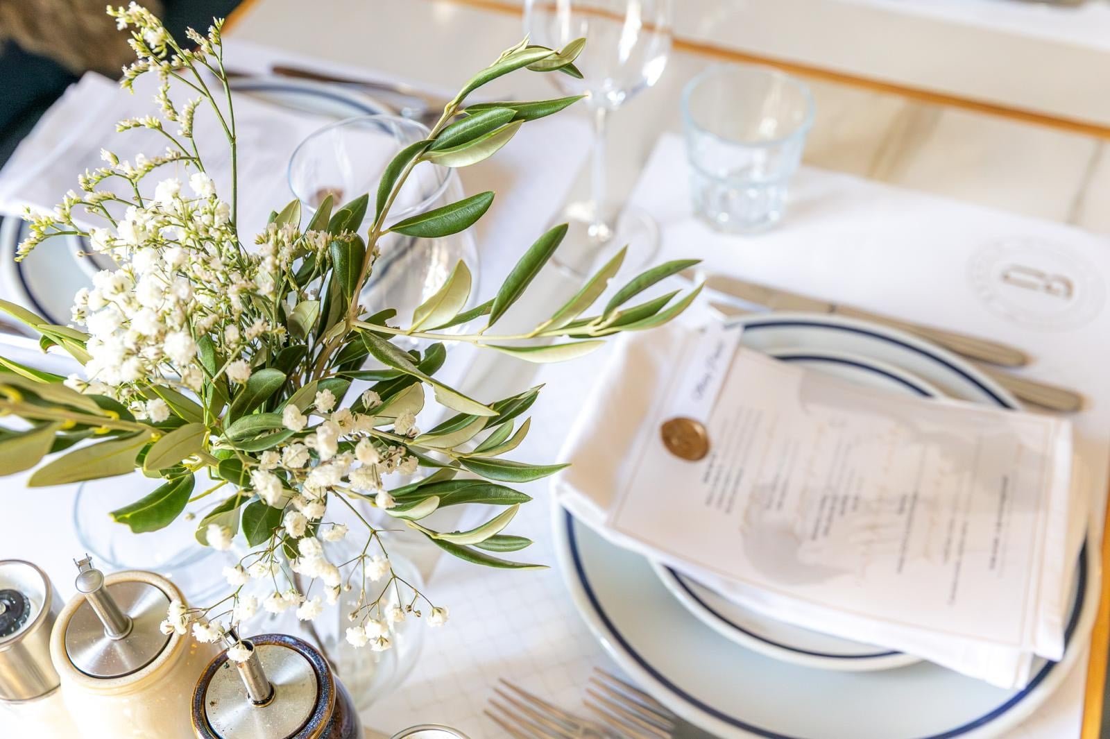 A Bronzin place setting with olive branches, flowers, printed menu, and white linen.